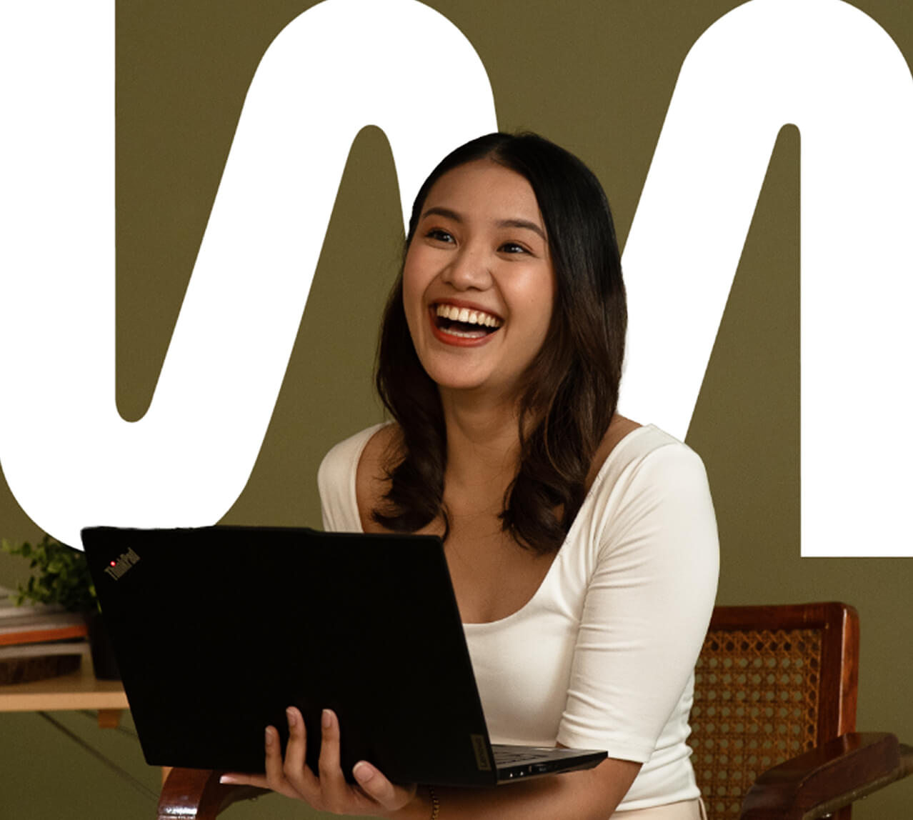 A woman sits on a chair, smiling and holding an open laptop, with a large white abstract design in the background.