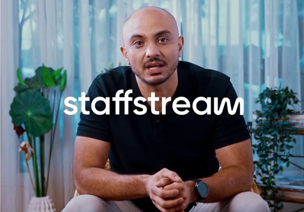 A man sits indoors with hands clasped, wearing a black shirt and watch, with the word "staffstream" overlaid in white text.