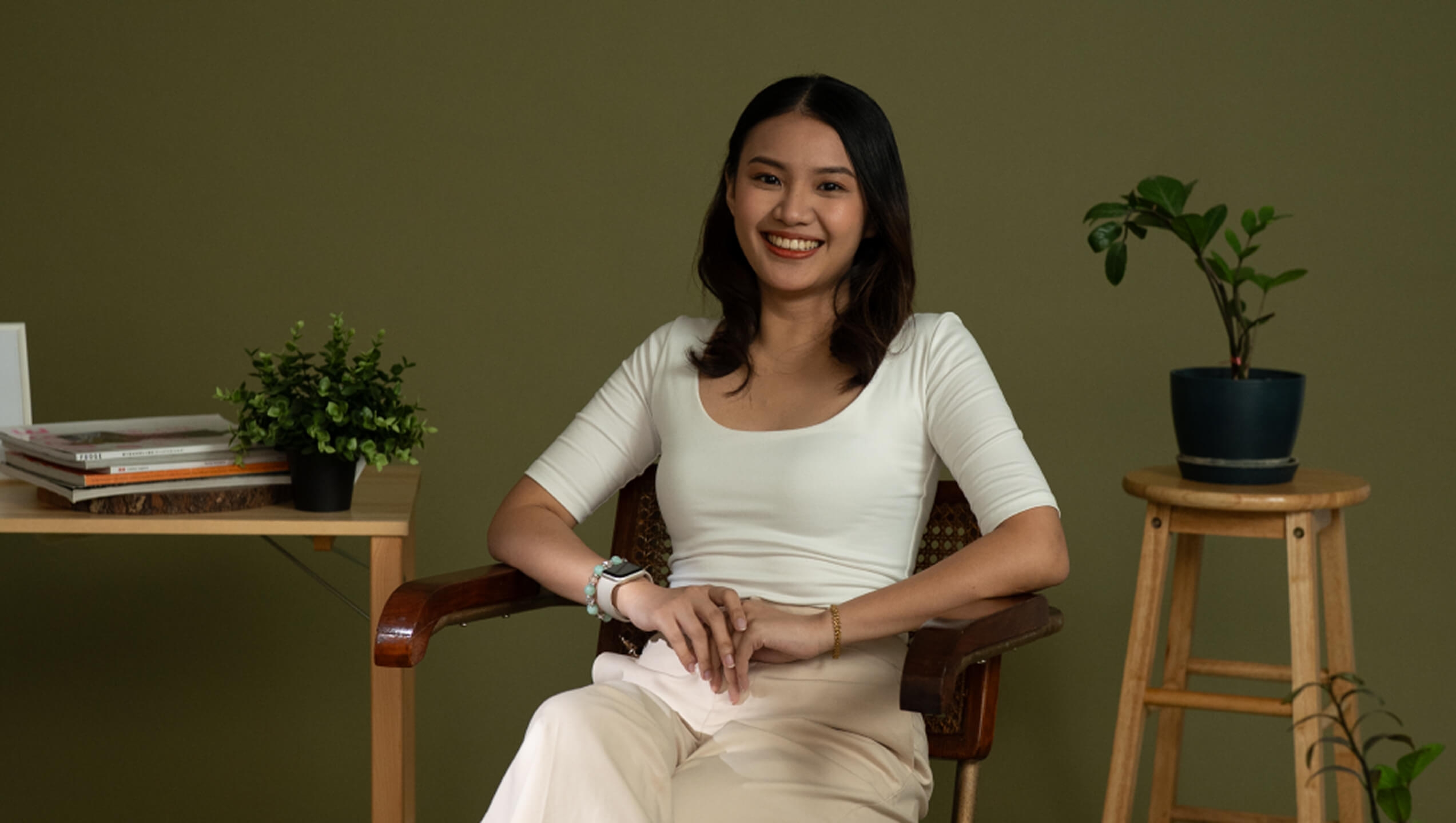 A woman sits and smiles in a wooden chair, surrounded by potted plants and a table with stacked books against a plain green wall.