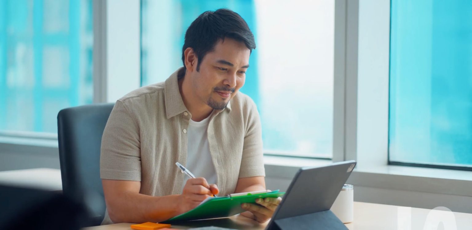A man sits at a desk with a clipboard, pen, and digital tablet, appearing focused while working in a bright office setting.
