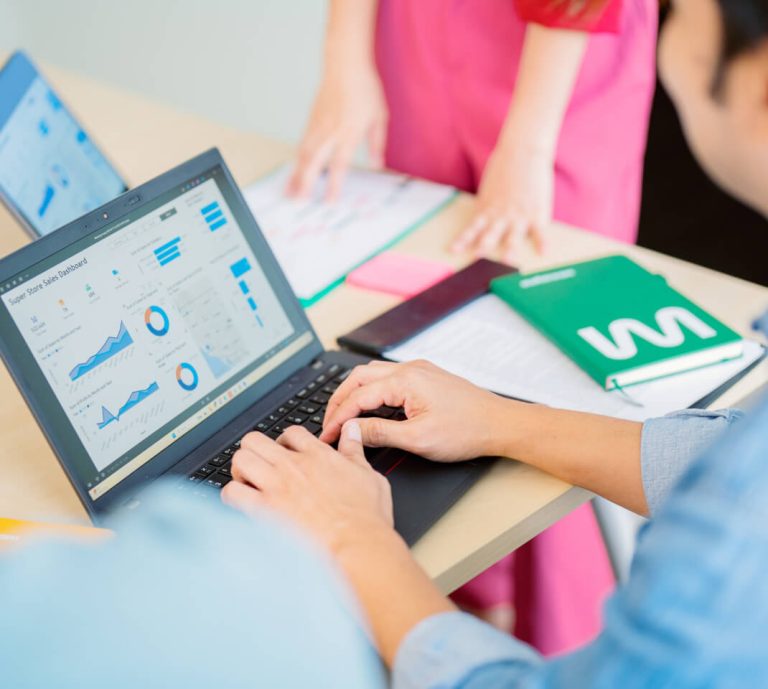 Person working on a laptop displaying data charts and graphs, with papers, notebooks, and another person standing nearby at a desk.