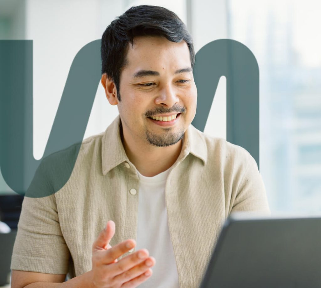 A man sits indoors, smiling and gesturing with his hand while looking at a laptop. Bold, semi-transparent letters overlay the image.