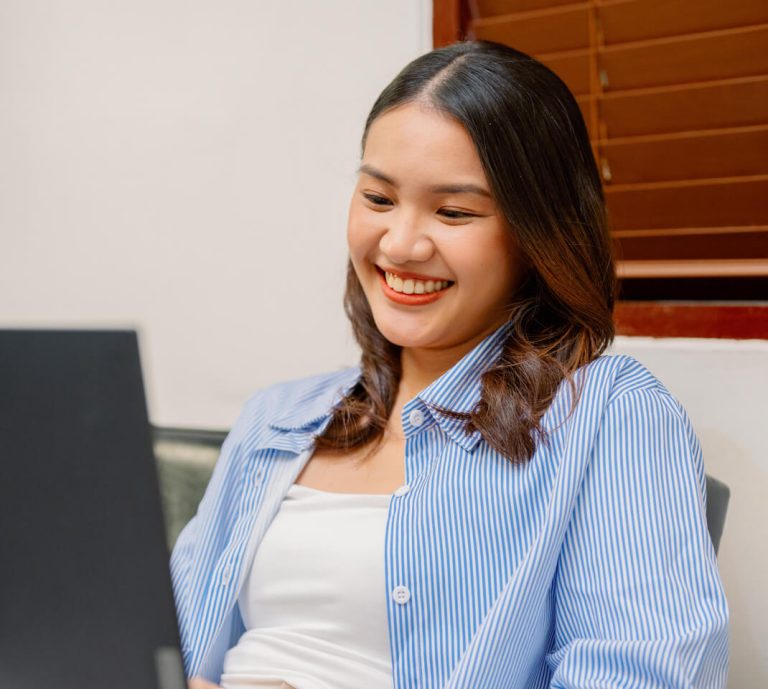 A woman in a blue and white striped shirt sits indoors, smiling while looking at a laptop screen.