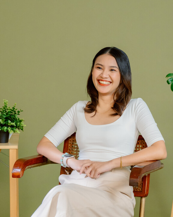 A woman with shoulder-length hair sits on a wooden chair, smiling, with her hands resting on her lap. There are green plants on a table and in the background.