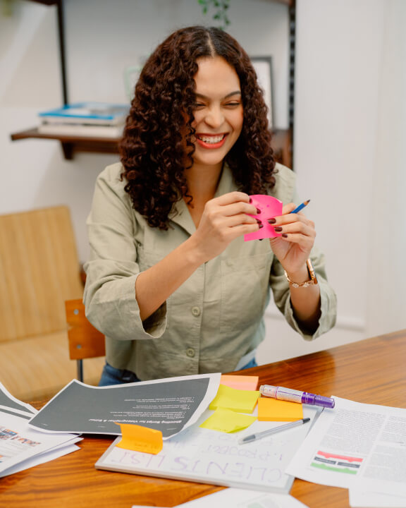 A woman sitting at a desk smiles while holding a pink sticky note. Papers, documents, and other sticky notes are spread out on the table in front of her.