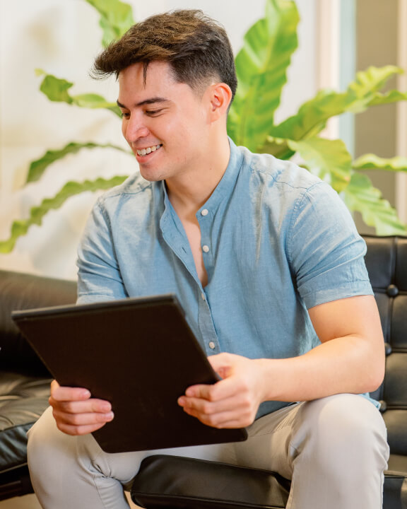 A man in a light blue shirt sits on a black sofa, smiling while holding a framed object, with a large green plant in the background.