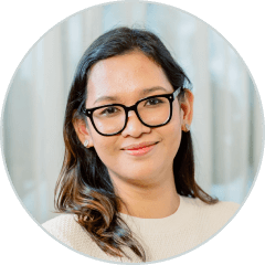 Woman with long brown hair and black glasses, wearing a light-colored top, smiling in front of a blurred light background.