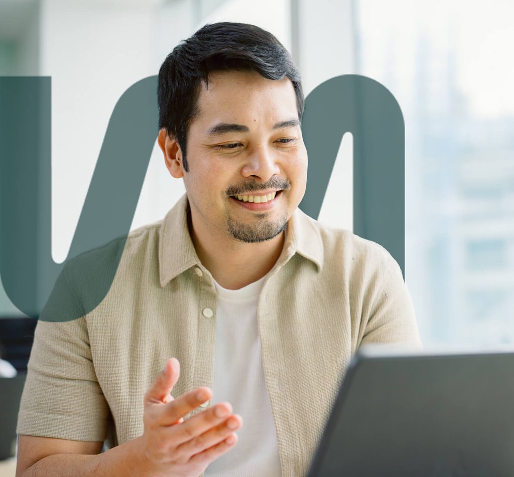 A man sits at a desk smiling and gesturing with one hand while looking at a laptop, with a large semi-transparent graphic overlay behind him.