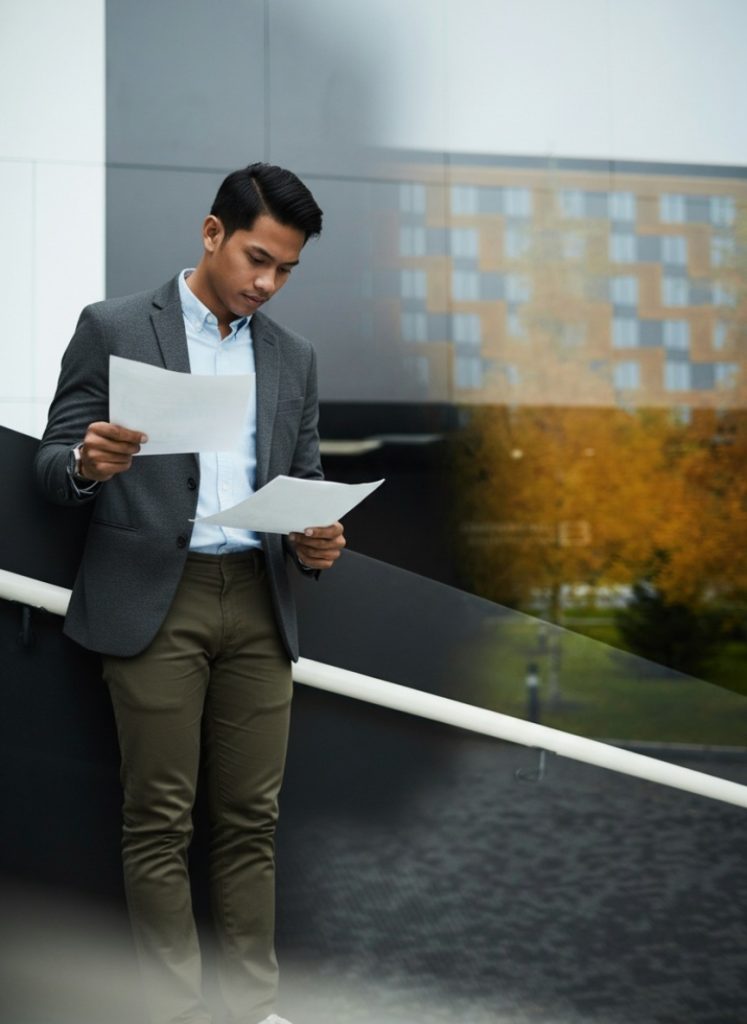 A man in business attire stands outdoors holding and reading two sheets of paper, with a modern building and trees in the background.