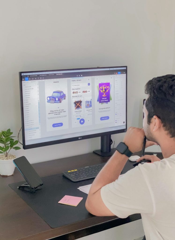 A person sits at a desk working on a computer displaying a user interface design app, with a phone, plant, and sticky notes also on the desk.