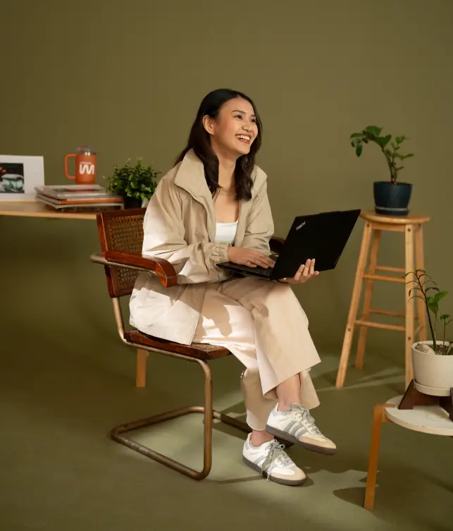 A woman sits on a chair holding a laptop and smiling. She is in a room with potted plants, a desk, a mug, and some books.