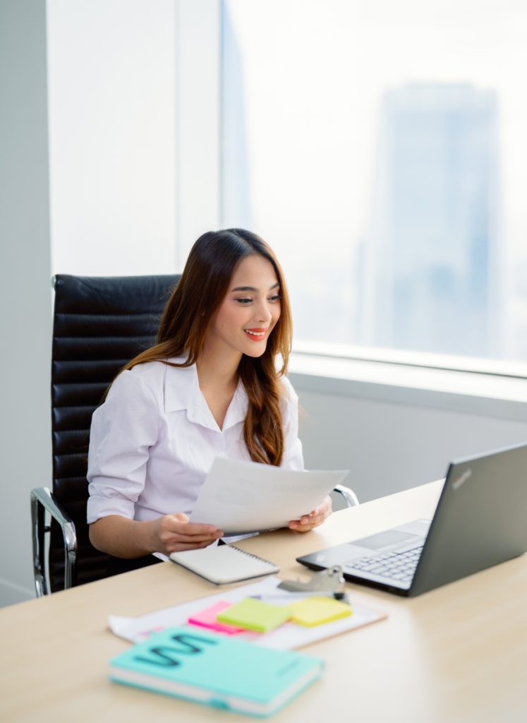 A woman sits at a desk with papers, notebooks, sticky notes, and a laptop, smiling while working in a modern office with large windows.