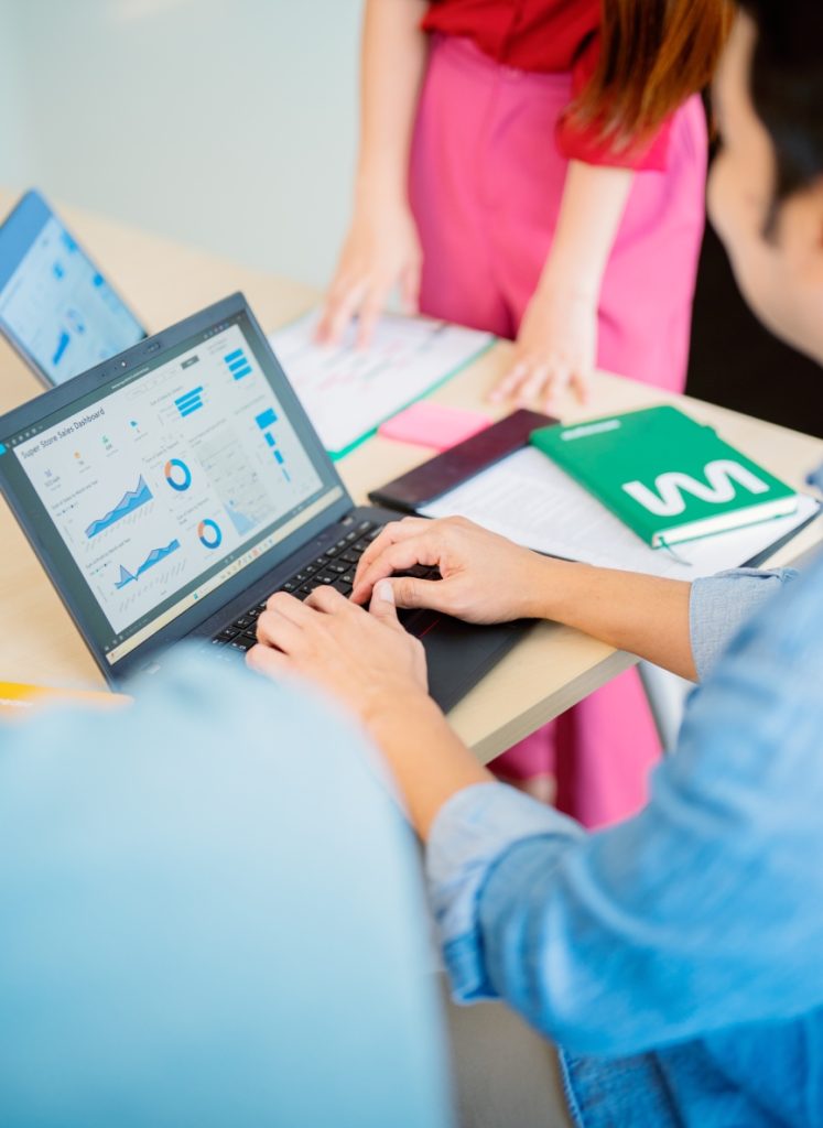 Two people work at a desk with laptops displaying graphs and charts, alongside notebooks and documents.