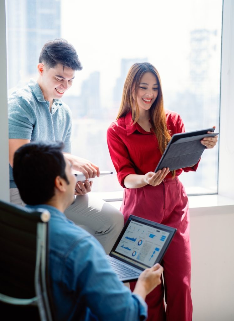 Three people in an office discuss data displayed on a tablet and a laptop near a large window with city buildings visible in the background.