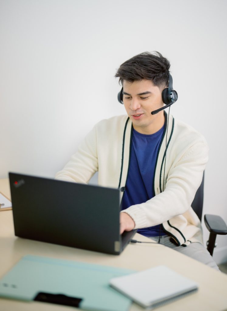 A person wearing a headset sits at a desk using a laptop, with notebooks and another device nearby.